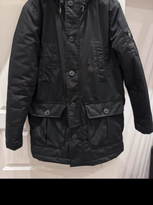 Svart parkajacka med knappar från clockhouse storlek L - Säljer en svart parkajacka med stora fickor och dragkedja på ärmen. Jackan har knyt i huvan och stängs med både knappar och dragkedja. Perfekt för kalla dagar och har en clean, enkel look som funkar till det mesta.