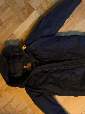 Marinblå Parajumpers vinterjacka storlek Young Small - 152/158  - Säljer en marinblå Parajumpers vinterjacka till kille/ junior från Masterpiece Series, modell right hand base. Jackan har flera fickor, huva och snygga orange detaljer. Quiltad insida och klassisk PJS-patch på ärmen. Perfekt för kalla dagar och riktigt stilren. Jackan är endast använd ett fåtal ggr av min son och är därmed i nyskick. Mer info kan du hitta på www.kidsbrandstore.se