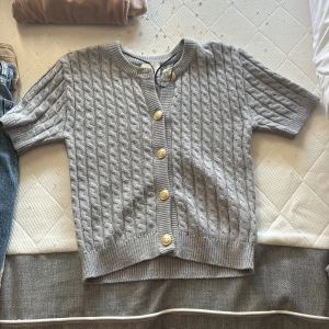 Grå kabelstickad kofta från H&M - Grå kabelstickad kofta från H&M i storlek XS. Koftan har korta ärmar, ribbade muddar och snygga guldknappar framtill. Perfekt för lager-på-lager och ger en klassisk vibe till din outfit.