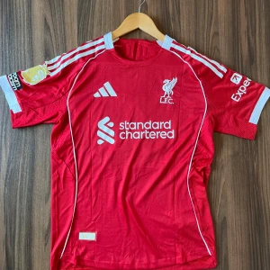 Liverpool Adidas fotbollströja Virgil M - Liverpool FC matchtröja från Adidas i rött med vita detaljer och tryck. Kortärmad modell i ventilerande polyester, med klubbmärke, sponsorloggor och namn/nummer Virgil 4 på ryggen. Slim fit passform och klassisk rund hals.