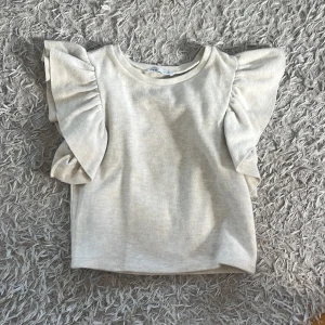 Beige topp från Zara  - Superfin beige topp från Zara med breda volangärmar som ger en riktigt snygg statement-look. Toppen är croppad och har en mjuk, ribbad känsla i bomullsmix. Perfekt för dig som vill ha något stilrent men ändå extra.