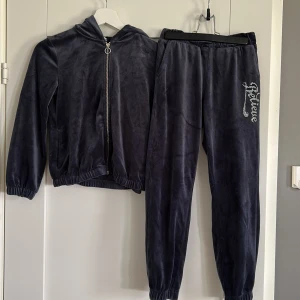 Mörkgråblå mjukisset, med tröja och byxor Lindex 134/140 - Mörkgråblå mjukisbyxor från Lindex i supermjuk velour med resår i midja och benslut. Snyggt strass-tryck med texten 'Believe' på ena benet. Matchande hoodie, med tragkedja och strass tryck på ryggen. Säljs som set