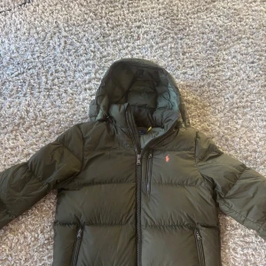 Grön dunjacka från Polo Ralph Lauren - Snygg mörkgrön dunjacka från Polo Ralph Lauren med huva och dragkedjor på fickorna. Jackan har en klassisk pufferstil och ikonisk röd logga på bröstet. Perfekt för kalla dagar och riktigt skön att bära. Fodrad och med hög krage för extra värme.