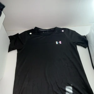 Svarta träningsshorts från Under Armour - Säljer ett par svarta träningsshorts och en T-shirt  från Under Armour med regnbågsfärgat logotryck på benet och framtill. Logotrycket lyser även nästan helt nytt.