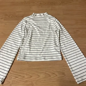 Vit och marinblå randig polotröja - Vit långärmad polotröja med tunna marinblå ränder från C. Tröjan har en klassisk polokrage och är tillverkad i mjukt material, perfekt för lager-på-lager. Enkel och stilren design som funkar till det mesta.