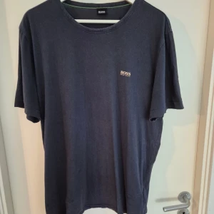 Mörkblå t-shirt från Hugo Boss - Snygg mörkblå t-shirt från Hugo Boss med diskret logga på bröstet och större logga på ryggen vid axeln. Klassisk rund hals och avslappnad passform. Perfekt för dig som gillar stilrena och enkla plagg med premiumkänsla.