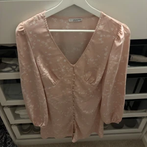 Ljusrosa blommig klänning Nelly - Supersöt ljusrosa klänning från Nelly med små vita blommor, v-ringning och knappar framtill. Långärmad modell i glansigt material som känns lyxigt och flowy. Perfekt för vår och sommar!