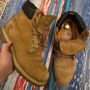 Timberland boots i beige skinn - Klassiska Timberland boots i beige skinn med robust platt sula och snörning. Skorna har svart vadderad kant upptill och tydlig Timberland-logga på sidan. Perfekta för dig som gillar streetstyle och vill ha en ikonisk look.