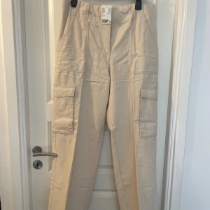 Beiga cargopants i bomull, strl 44 - Snygga beiga cargopants med hög midja och raka ben. Byxorna har stora fickor på sidorna och bak, vilket ger en cool och avslappnad look. Tillverkade i bomull för en skön känsla och passar perfekt till streetwear-stilen.