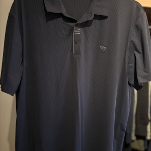 Mörkblå pikétröja från Armani - Snygg mörkblå pikétröja från Armani med diskreta ränder och klassisk krage. Tröjan har korta ärmar, knappslå framtill och den ikoniska Armani-loggan broderad på bröstet. Perfekt för en stilren och avslappnad look.