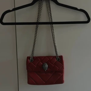 Röd quiltad axelväska från Kurt Geiger - Säljer en snygg röd axelväska från Kurt Geiger i quiltat skinn. Väskan har en kraftig silverfärgad kedja och ett coolt metallspänne framtill. Perfekt storlek för det viktigaste och ger en riktigt trendig vibe till din outfit.