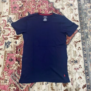 Mörkblå t-shirt från Polo Ralph Lauren - Snygg mörkblå t-shirt från Polo Ralph Lauren med klassisk rund hals och korta ärmar. T-shirten har en liten röd broderad logga vid nederkanten och en diskret röd detalj på insidan av halsen. Tillverkad i mjuk bomull för skön känsla.
