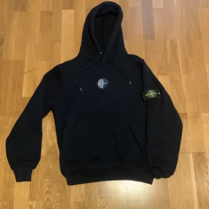 Svart hoodie från Stone Island - Svart hoodie från Stone Island med klassisk kompasspatch på vänster ärm och broderad logga på bröstet. Tröjan har huva med snörning, känguruficka och ribbade muddar. Perfekt för dig som gillar streetwear och vill ha en clean look.