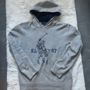Grå hoodie från Polo Ralph Lauren - Snygg grå hoodie från Polo Ralph Lauren med stor blå broderad polospelare och RL 67 på bröstet. Tröjan har huva med mörkblå insida, ribbade muddar och är tillverkad i mjuk bomull. Perfekt för chill dagar och sportig stil.