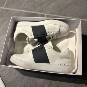 Valentino Garavani Open sneakers vit/svart - Säljer ett par snygga Valentino Garavani Open sneakers i vitt skinn med bred svart rem över mitten. Skorna har klassisk rund tå, vita snören och ikoniska nitar på hälen. Perfekta för dig som vill ha en clean men lyxig look. Har knappt använt dem eftersom dem var för små. Känner ni sug på dem är det bara höra av er. Mvh Arian
