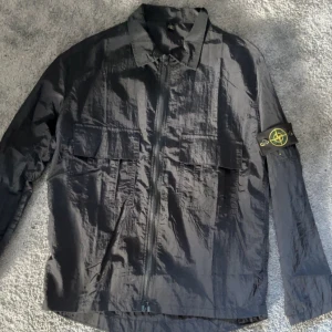 Svart overshirt från Stone Island - Svart overshirt från Stone Island med klassisk patch på vänster ärm. Jackan har två stora bröstfickor med lock, dragkedja framtill och krage. Materialet är glansigt och lätt, perfekt för lager-på-lager. Långärmad och stilren design som funkar året runt.