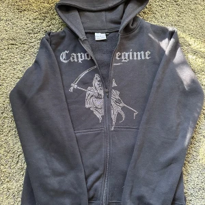 Svart rihstone zip up hoodie - Svart hoodie från Capo Regime med dragkedja och huva. Framsidan har ett stort tryck med dödskallemotiv och text i silvriga rihstone detaljer. Tröjan är långärmad och har en klassisk passform. Perfekt för dig som gillar streetwear och edgy stil.