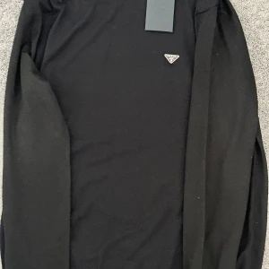 Svart stickad tröja från Prada - Svart stickad tröja från Prada med rund halsringning och diskret logga på bröstet. Tröjan har långa ärmar och ribbade muddar vid hals, ärmslut och nederkant. Perfekt för dig som gillar stilren och lyxig design.