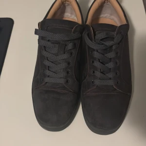 Christian Louboutin svarta sneakers - Snygga svarta sneakers från Christian Louboutin med klassisk röd sula. Skorna har rund tå, platt sula och är grått i mocka med svarta snören. Insidan är beige och de har en stilren, minimalistisk design som passar perfekt till streetwear.