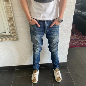 Tiger of Sweden Jeans - Snygga blå jeans från Tiger of Sweden i modellen Evolve, tillverkade i Italien. Jeansen har en klassisk femficksdesign, orangea sömmar och en tvättad look med slitningar. Passformen är smal och benen är raka. Perfekta för dig som gillar en modern och avslappnad stil.