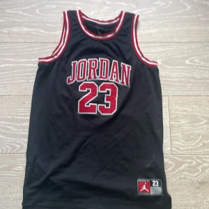 Jordan basketlinne svart röd nr 23 - Säljer ett svart basketlinne från Jordan med röd och vit kant vid hals och ärmhål. Stort tryck med JORDAN och nummer 23 på bröstet samt på ryggen. Tillverkat i meshmaterial som andas, perfekt för sport eller streetwear.