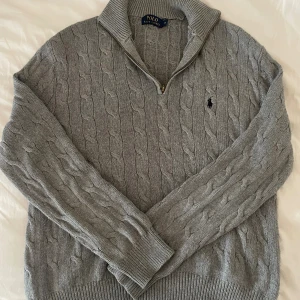 Grå kabelstickad tröja Polo Ralph Lauren - Kabelstickad grå tröja från Polo Ralph Lauren med halv dragkedja och klassisk broderad logga på bröstet. Tröjan har hög krage, ribbade muddar och är långärmad. Perfekt för lager-på-lager och snygg till jeans eller chinos.