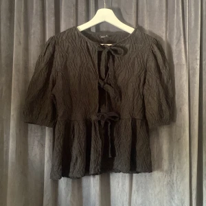 Svart blus med knyt från Gina Tricot - Svart blus från Gina Tricot med vågig struktur i tyget och tre knyt framtill. Modellen har trekvartsärmar och en lätt volang nertill. Tillverkad i polyester och elastan för en skön och följsam känsla. Perfekt för dig som gillar unika detaljer och en luftig look.