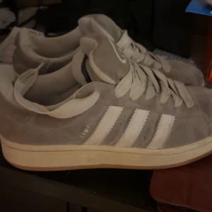 Grå Adidas Campus  - Snygga grå Adidas Campus sneakers i mocka med klassiska vita ränder på sidorna och vit sula. Skorna har rund tå och platt sula, samt breda vita skosnören. Perfekta för en avslappnad och stilren look.