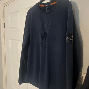 Marinblå Sail Racing sweatshirt - Snygg marinblå sweatshirt från Sail Racing med broderad logga och text på bröstet samt patch på ärmen. Tröjan har rund hals, långa ärmar och en relaxed passform. Perfekt för dig som gillar stilren design och sportiga detaljer.