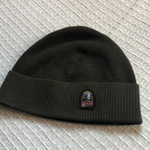 Mörkgrön mössa från Parajumpers - Snygg mörkgrön mössa från Parajumpers med ribbad kant och broderad patch framtill med texten 'PJS Anchorage'. Mössan är stickad i en blandning av ull och akryl, perfekt för kalla dagar. Klassisk och enkel design som funkar till de flesta stilar.