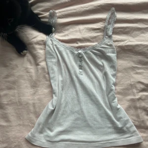 Vit ribbad linnetopp med spetsdetaljer - Supersöt vit ribbad linnetopp med spetskant runt urringningen och axelbanden. Dekorerad med små knappar framtill för extra detalj. Perfekt att styla med jeans eller kjol för en fräsch look.