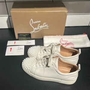 Christian Louboutin vita sneakers med nitar - Säljer ett par vita sneakers från Christian Louboutin i skinn med ikoniska röda sulor och coola nitar över tån. Skorna har snörning och en klassisk låg siluett. Kommer med originalkartong, dustbag och äkthetskort. Perfekta för dig som vill sticka ut med lyxig streetstyle.