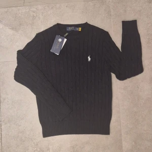 Mörkblå stickad tröja från Polo Ralph Lauren - Svart kabelstickad tröja från Polo Ralph Lauren Klassisk stickad tröja i mörk       marinblå/svart nyans med vit broderad logga på bröstet.                                                                                                 Storlek: M, men passar till XS                                                                              Skick: Helt ny med tag kvar.  Passar perfekt till både vardag och mer uppklätt. Jag är storlek L på bilden.