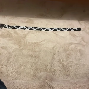 Svartvitt randigt flätat bälte - Snyggt flätat bälte i svart och vitt med randigt mönster. Bältet har detaljer i svart skinn vid spännet och änden, samt ett klassiskt metallspänne. Perfekt accessoar för att lyfta din outfit med en cool kontrast.