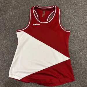 Röd och vit linnetopp från Wilson - Sportig linnetopp från Wilson i rött och vitt med coolt diagonalt mönster. Toppen har brottarrygg och kontrastfärgade kanter vid hals och ärmhål. Perfekt för dig som gillar en aktiv stil och vill sticka ut på tennisbanan.