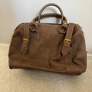 Brun axelväska i skinnlook - Stilren brun axelväska med snygga guldfärgade metalldetaljer och dubbla handtag. Väskan har en klassisk form, dragkedja upptill och dekorativa spännen framtill. 