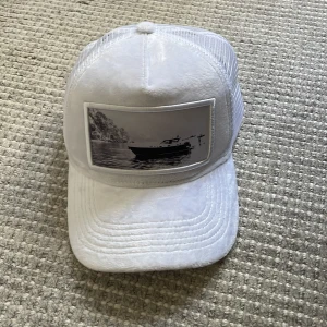 Vit keps från Emilio Pendenza - Snygg vit keps från Emilio Pendenza med mesh på sidorna och baksidan för extra ventilation. Framtill finns en svartvit bild av en båt på vattnet som ger kepsen en unik vibe. Justerbar passform med plastspänne bak. Perfekt för dig som gillar stilrena accessoarer. Använd fåtal gånger. Skriv vid minsta funderingar 