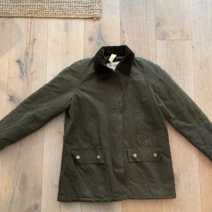 Olivgrön parkajacka från Brixtol S - Snygg olivgrön parkajacka från Brixtol Textiles med quiltat foder, krage och muddar i manchester. Jackan har två stora fickor med knappar och dragkedja framtill. Perfekt för höst och vinter när du vill ha både stil och funktion. Använd endast ett fåtal gånger.