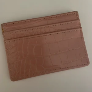 Rosa plånbok i croco-imitation från Glitter - Slimmad rosa plånbok med croco-mönster och flera kortfack. Snygg och stilren design i syntetmaterial, perfekt för att hålla koll på korten i fickan eller väskan. Enkel att matcha med olika stilar och outfits.