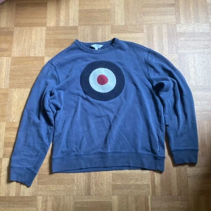 Blå sweatshirt Ben Sherman med target - Snygg blå sweatshirt från Ben Sherman i bomull, med ett stort broderat target-märke i svart, beige och rött på bröstet. Klassisk rund hals och långa ärmar, perfekt för en chill och stilren look. Vintage original!!
