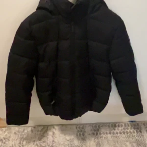 Svart pufferjacka från FB Sister - Säljer en svart pufferjacka från FB Sister i storlek M. Jackan har luva, dragkedja framtill och quiltad design med breda kanaler. Perfekt för kalla dagar och riktigt skön att bära. Snygg och enkel att matcha med det mesta. Funkar även till tjejer och  killar. Ny i stick använd bara 2 gånger.