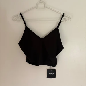 Svart croppad linnetopp från SHEIN - En svart croppad linnetopp från SHEIN med smala axelband och v-ringad hals. Toppen är enkel och stilren, perfekt att matcha med jeans eller kjol. Tillverkad i mjuk polyesterblandning med lite stretch för en bekväm passform.