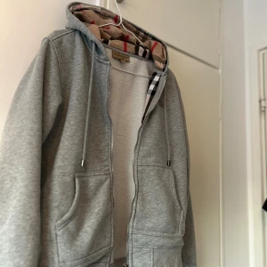 Grå hoodie från Burberry med dragkedja - Grå hoodie från Burberry med klassiskt rutigt foder i huvan och broderad logga på bröstet. Tröjan har dragkedja framtill, snörning i huvan och två stora fickor. Perfekt för dig som gillar stilren design med en touch av lyx. Priset kan diskuteras 