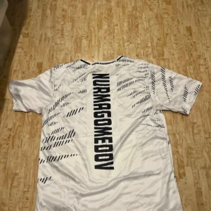 UFC Nurmagomedov t-shirt Venum vit - Säljer en vit UFC t-shirt från Venum med Nurmagomedov-tryck på ryggen. T-shirten har korta ärmar, svart mönster och blåa detaljer med crypto.com-logga på framsidan. Perfekt för dig som gillar MMA och sportig stil.