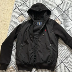 Svart hoodiejacka från Polo Ralph Lauren - Svart hoodiejacka från Polo Ralph Lauren med dragkedja, huva med snörning och två fickor med dragkedja. Jackan har röd broderad logga på bröstet och ribbade muddar. Materialmix av bomull och polyester för en skön känsla.