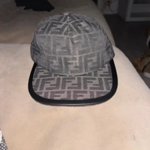 Grå Fendi keps med FF-mönster - Snygg grå keps från Fendi med det klassiska FF-logomönstret över hela kepsen. Skärmen har en svart kant och kepsen är justerbar baktill. Materialet är bomull och polyester, vilket gör den både bekväm och stilren. Perfekt för dig som vill ha en exklusiv streetwear-look.