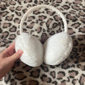 Vita fluffiga öronmuffar - Supermjuka och fluffiga vita öronmuffar som håller dig varm under kalla dagar. De har en klassisk rund form och ett brett band som sitter bekvämt över huvudet. Perfekt accessoar för vintern och ger en söt touch till din outfit.