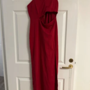 Röd långklänning med oneshoulder - Snygg röd långklänning med oneshoulder och smala axelband. Klänningen har en stilren design med en liten slits nertill och cutout-detalj vid midjan. Perfekt för dig som vill sticka ut med en elegant och modern look. Den är helt ny och kommer från rök- och djurfritt hem. Titta på bilderna för mer info. 