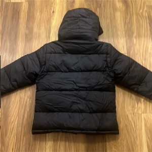 Svart pufferjacka från Burberry - Svart pufferjacka från Burberry med huva och klassiskt rutigt foder. Jackan har vita Burberry London England-broderier på bröstet, dragkedjor på fickorna och tryckknappar framtill. Perfekt för kalla dagar med sin tjocka, quiltade design och snygga detaljer.