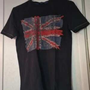 Svart t-shirt med Union Jack-tryck - Svart t-shirt med korta ärmar och ett stort, slitet Union Jack-tryck i rött, vitt och blått på bröstet. T-shirten har synliga kontrastsömmar längs ärmslut och nederkant. I nederkanten kan man även se tecken på slitage. Skön passform och klassisk rund halsringning.